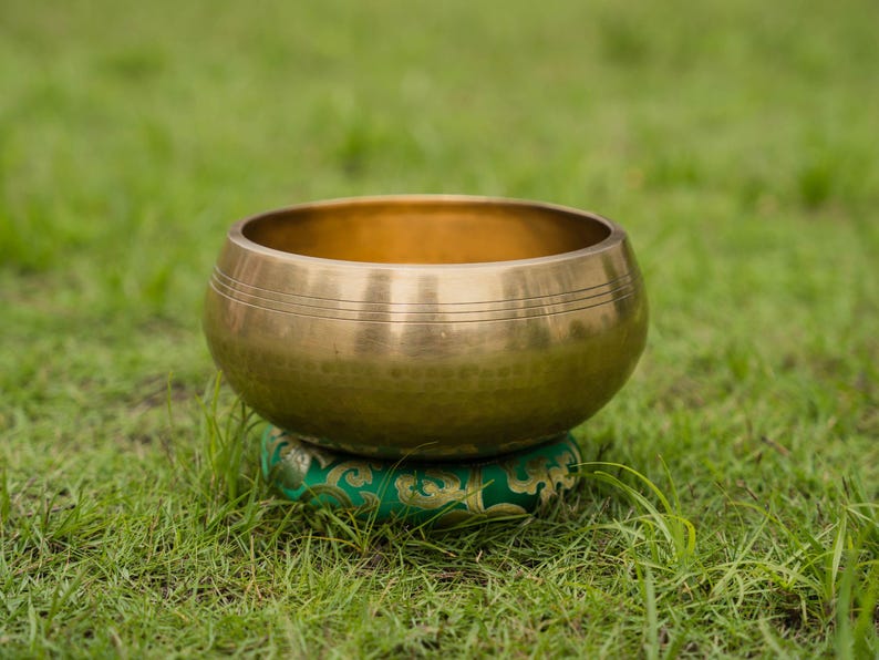 May include: A golden singing bowl with etched lines rests on a green patterned cushion, set against a backdrop of green grass. The bowl's metallic surface reflects the light, highlighting its rounded shape.