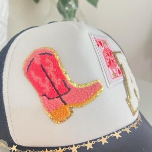 May include: A black and white trucker hat with a white front panel. The hat features a pink cowboy boot patch, a playing card patch, and a gold star chain along the brim. The letter "A" is also on the hat.