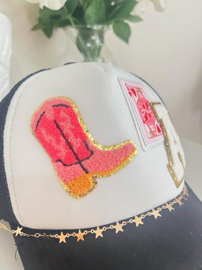 May include: A black and white trucker hat with a white front panel. The hat features a pink cowboy boot patch, a playing card patch, and a gold star chain along the brim. The letter "A" is also on the hat.