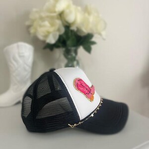 May include: Black and white trucker hat with a pink and orange design on the front. The hat has a black mesh back, a white front panel, and a black brim. A gold star chain is attached to the brim.