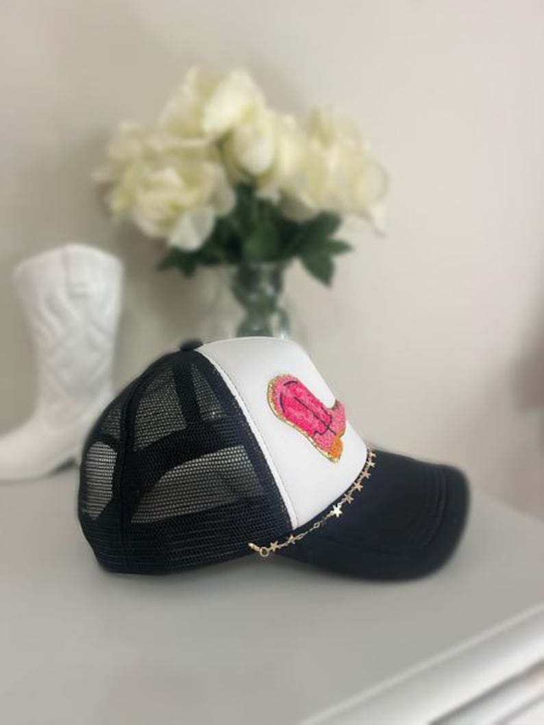 May include: Black and white trucker hat with a pink and orange design on the front. The hat has a black mesh back, a white front panel, and a black brim. A gold star chain is attached to the brim.