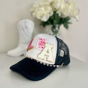 May include: A black and white trucker hat with the letters "LA" and a pink heart design. The hat has a gold star chain along the brim and a black brim. A white cowboy boot is in the background.