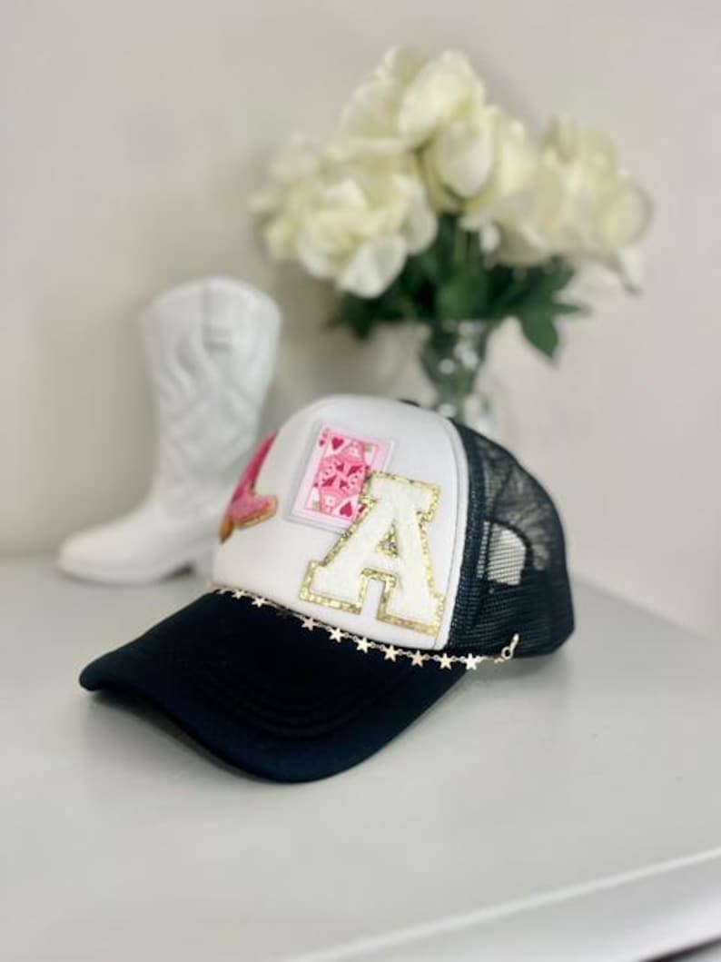May include: A black and white trucker hat with the letters "LA" and a pink heart design. The hat has a gold star chain along the brim and a black brim. A white cowboy boot is in the background.