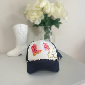 May include: A navy blue and white trucker hat with the letters "LA" and a pink cowboy boot patch. The hat has a gold star chain around the brim and a mesh back. A white cowboy boot and a bouquet of white flowers are in the background.