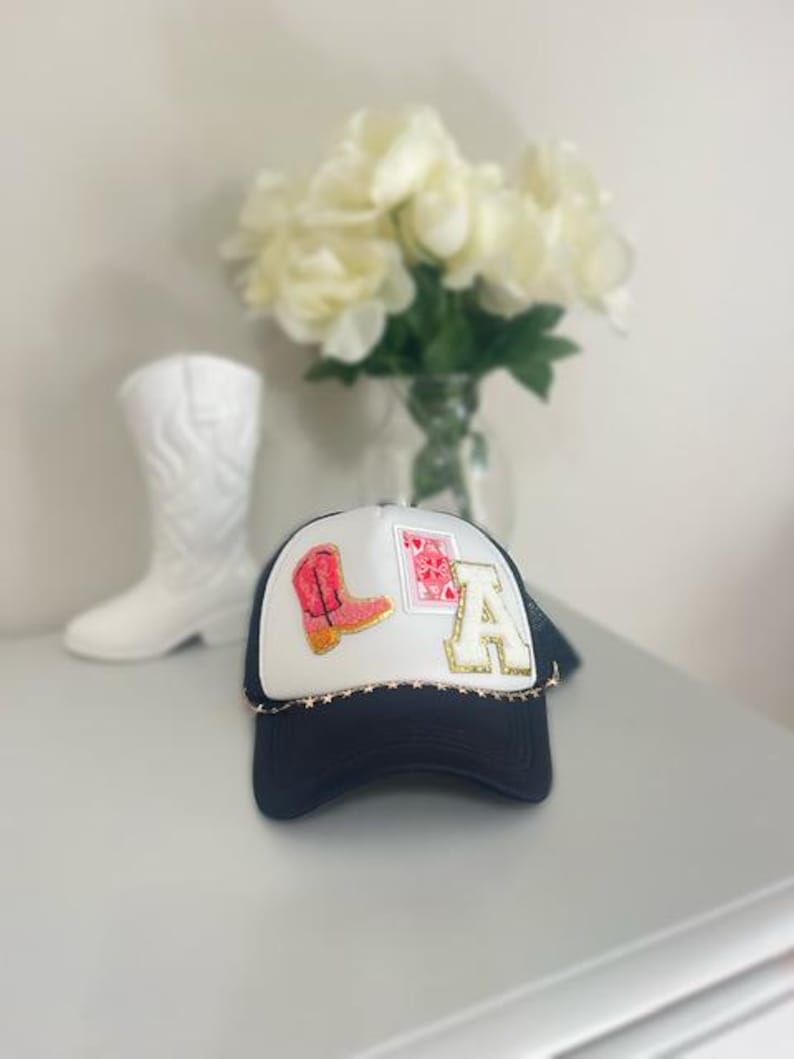 May include: A navy blue and white trucker hat with the letters "LA" and a pink cowboy boot patch. The hat has a gold star chain around the brim and a mesh back. A white cowboy boot and a bouquet of white flowers are in the background.