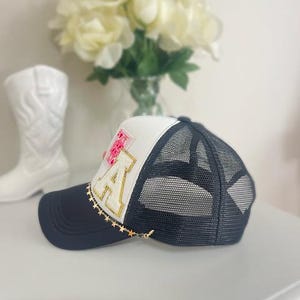 May include: A navy blue and white trucker hat with a mesh back. The hat features a gold chain with star charms and a patch with the letter "A" and floral accents. A white cowboy boot and flowers are in the background.