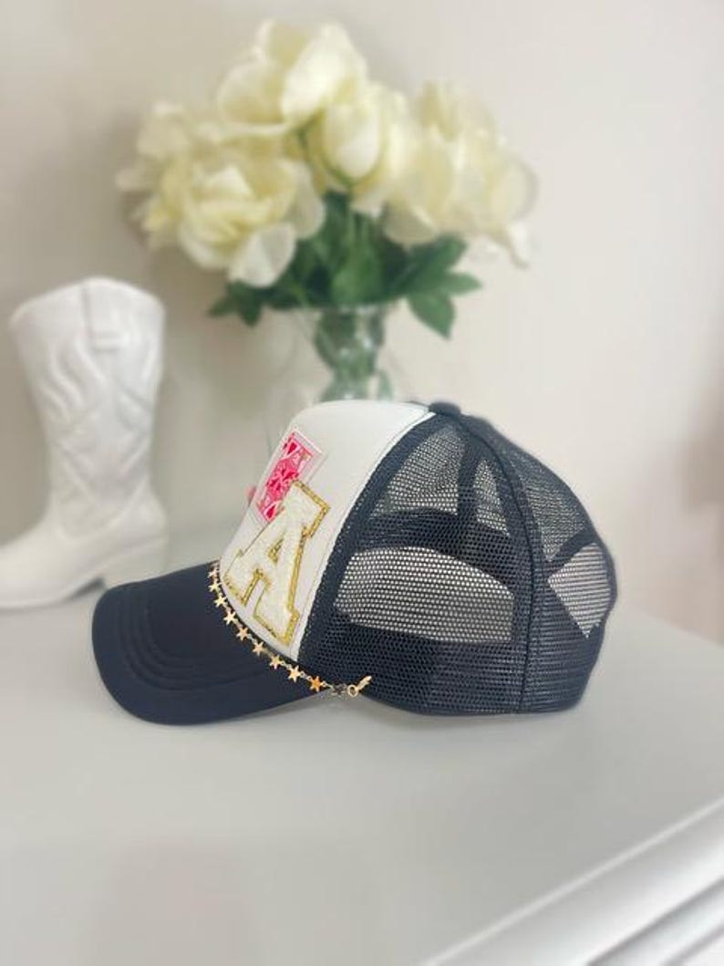 May include: A navy blue and white trucker hat with a mesh back. The hat features a gold chain with star charms and a patch with the letter "A" and floral accents. A white cowboy boot and flowers are in the background.
