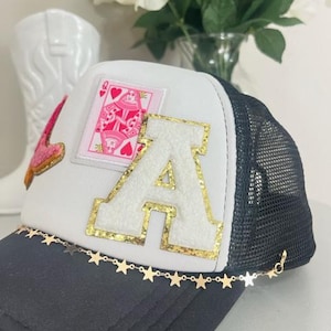 May include: A trucker hat with a white front panel, black mesh back, and a curved brim. The hat features a large gold-trimmed letter "A", a pink playing card patch, and a gold star chain along the brim.