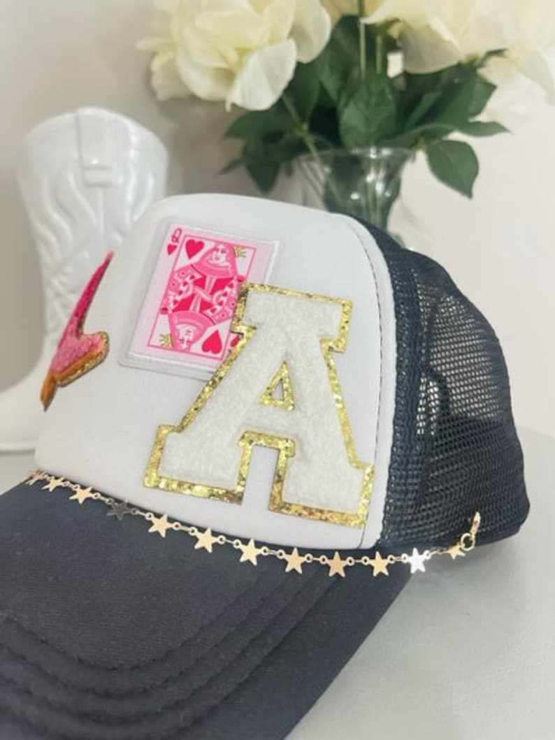 May include: A trucker hat with a white front panel, black mesh back, and a curved brim. The hat features a large gold-trimmed letter "A", a pink playing card patch, and a gold star chain along the brim.