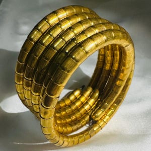 Puede incluir: Una pulsera dorada enrollada, con un diseño segmentado y texturizado. La pulsera se enrolla varias veces, creando un efecto en capas. La joya está hecha de un material metálico brillante.