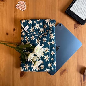 Könnte beinhalten: Eine tealfarbene, gesteppte Tablethülle mit weißem Gänseblümchenmuster, neben einem blauen Tablet und einem Kindle. Ein Zweig weißer Blumen liegt auf der Hülle. Weitere Gegenstände auf der Holzoberfläche sind ein Bücheraufkleber, Lipgloss und eine Kerze.
