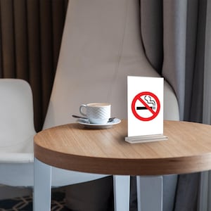 Könnte beinhalten: Ein kleiner, runder Holztisch mit weißem Fuß trägt eine weiße Kaffeetasse und ein "Rauchen verboten"-Schild. Das Schild ist weiß mit einem roten Kreis und einer Grafik einer durchgestrichenen Zigarette. Im Hintergrund ist ein weißer Stuhl zu sehen.