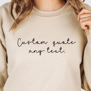 May include: A beige sweatshirt with the text "Custom quote any text" in black script. The crew neck sweatshirt is a neutral color and has long sleeves. The text is centered on the front of the shirt.