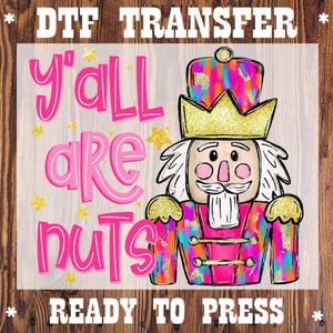 May include: A DTF transfer featuring a colorful nutcracker design with the text "y'all are nuts." The design includes gold stars and the words "READY TO PRESS" in a bold font. The nutcracker wears a gold crown and a pink jacket.