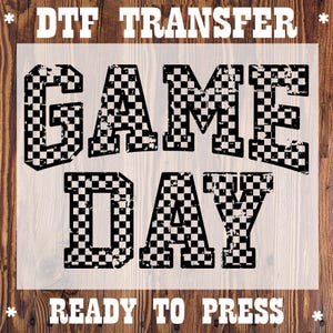 May include: DTF Transfer with the words "GAME DAY" in large, distressed, checkered letters. The words "READY TO PRESS" are at the bottom. The design is on a clear background, suitable for heat transfer applications.