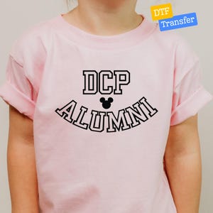 May include: A light pink short-sleeved t-shirt featuring a black graphic print on the chest. The text reads DCP in a block font above a small black mouse ear silhouette, with the word ALUMNI arched underneath in a matching black block font.