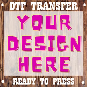 May include: DTF transfer with the words "YOUR DESIGN HERE" in bright pink, outlined in yellow. The words "DTF TRANSFER" and "READY TO PRESS" are above and below the design in white. The background is a wood grain.