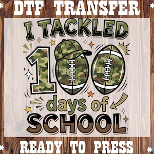 May include: A DTF transfer design with the text "I TACKLED 100 days of SCHOOL" in black and brown. The design includes a football, a hat, and the number 100 in a camouflage pattern. The words "READY TO PRESS" are at the bottom.