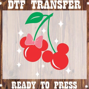 May include: A DTF TRANSFER READY TO PRESS graphic featuring two red cherry-shaped silhouettes with mouse-ear outlines, a pink bow on one cherry, a green stem with a leaf, and white star accents against a light background with wood grain.