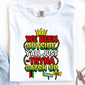 May include: White t-shirt with the text "WE BEEN OUTSIDE Y'ALL JUST -TRYNA CATCH UP" in red, yellow, and green, with a gold crown and chain design. Includes "FREEDOM" and "1865" signs.