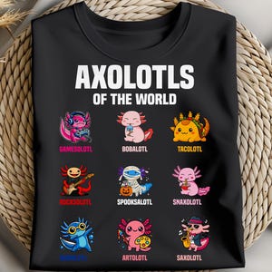 May include: Black t-shirt featuring the text "AXOLOTLS OF THE WORLD" above nine cartoon axolotl characters. Each axolotl is uniquely designed, with names like "GAMESOLOTL" and "TACOLOTL" below them. The shirt is displayed on a woven background.