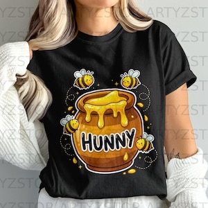May include: A black short-sleeved t-shirt featuring a graphic of a brown honey pot with dripping yellow honey and the word HUNNY printed in black. Four cartoon bees with yellow and black stripes and white wings surround the honey pot.