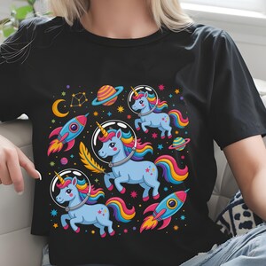 May include: Black t-shirt featuring a colorful graphic of space-themed unicorns. The design includes blue unicorns with rainbow manes and tails, rockets, planets, stars, and a crescent moon. The unicorns are wearing astronaut helmets.