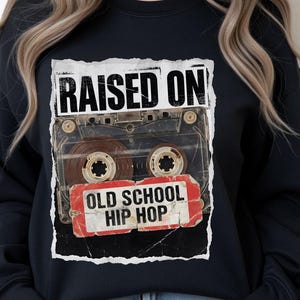 May include: Black sweatshirt featuring a graphic of a cassette tape with the words "RAISED ON" above and "OLD SCHOOL HIP HOP" below. The design has a distressed, vintage aesthetic.