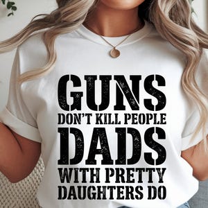 May include: White t-shirt with the text "GUNS DON'T KILL PEOPLE DADS WITH PRETTY DAUGHTERS DO" in a bold, distressed black font. The shirt has short sleeves and a crew neck.
