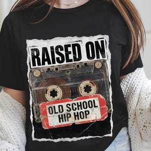 May include: Black t-shirt featuring a distressed graphic of a cassette tape with the words "RAISED ON" above and "OLD SCHOOL HIP HOP" below. The design is in white and red, evoking a vintage aesthetic.