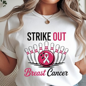 May include: White t-shirt with a bowling-themed graphic. The design features bowling pins and a pink bowling ball with a breast cancer awareness ribbon. Text reads "Strike Out Breast Cancer" in black and pink.