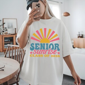 Puede incluir: Camiseta blanca con un dise&ntilde;o gr&aacute;fico colorido que presenta un sol y las palabras "SENIOR sunrise CLASS OF 2026" en azul, rosa y amarillo. La camiseta es de estilo casual y oversize.