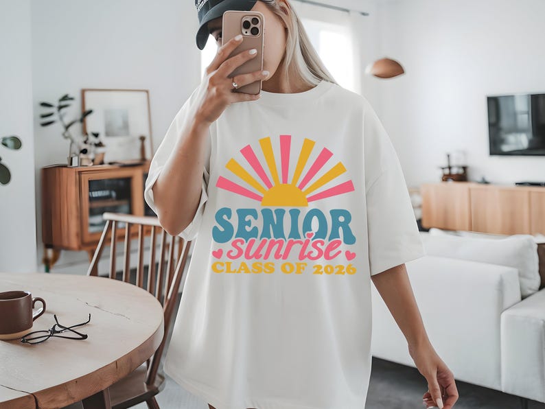 Puede incluir: Camiseta blanca con un dise&ntilde;o gr&aacute;fico colorido que presenta un sol y las palabras "SENIOR sunrise CLASS OF 2026" en azul, rosa y amarillo. La camiseta es de estilo casual y oversize.
