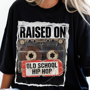 May include: Black t-shirt featuring a distressed graphic of a cassette tape with the words "RAISED ON" above and "OLD SCHOOL HIP HOP" below. The design is printed on a black, short-sleeved shirt.