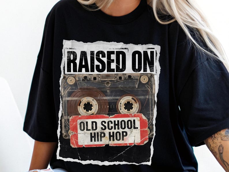 May include: Black t-shirt featuring a distressed graphic of a cassette tape with the words "RAISED ON" above and "OLD SCHOOL HIP HOP" below. The design is printed on a black, short-sleeved shirt.