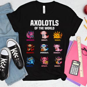 May include: Black t-shirt with the text "AXOLOTLS OF THE WORLD" and cartoon axolotl characters. The axolotls are depicted in various styles, including a gamer, a musician, and a taco-themed axolotl. The shirt is paired with jeans and black sneakers.