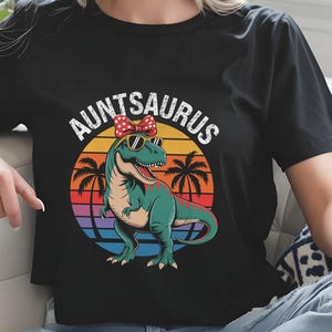 Op de afbeelding: Zwart t-shirt met een kleurrijke afbeelding van een dinosaurus met zonnebril en strik, met de tekst "Auntsaurus" erboven. De dinosaurus is groen met een rode mond, tegen een zonsondergang met palmbomen.