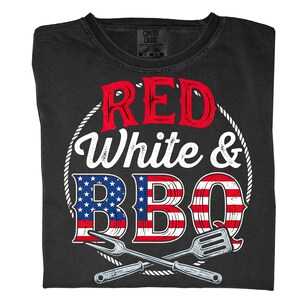 May include: Black t-shirt with the words "RED White & BBQ" in red and white lettering. The letters "B" and "O" are designed with red and white stripes, and the letter "B" has a blue background with white stars. Crossed grilling tools are at the bottom.