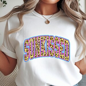 以下が含まれることがあります： 大胆なアーチ型デザインで「SUNKISSED」の文字が入った白いTシャツ。テキストは、黄色の背景、ピンクの輪郭、水色の内側の境界線を持つヒョウ柄のパターンが特徴です。 シャツは袖をまくっています。