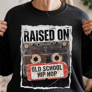 May include: Black sweatshirt featuring a graphic of a cassette tape with the words "RAISED ON" above and "OLD SCHOOL HIP HOP" below. The cassette tape is brown and black with red and white accents. The sweatshirt is a crew neck.