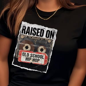 May include: Black t-shirt featuring a cassette tape graphic with the words "RAISED ON OLD SCHOOL HIP HOP" in a distressed style. The design includes a vintage cassette tape image, evoking a retro music theme.