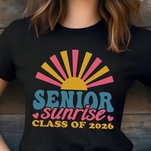 Puede incluir: Camiseta negra con un dise&ntilde;o retro. Las palabras "SENIOR sunrise" est&aacute;n en verde azulado y rosa, con un gr&aacute;fico de sol amarillo y rosa. El texto "CLASS OF 2026" es amarillo. Un dise&ntilde;o colorido para la graduaci&oacute;n.