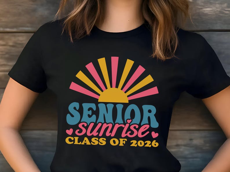 Puede incluir: Camiseta negra con un dise&ntilde;o retro. Las palabras "SENIOR sunrise" est&aacute;n en verde azulado y rosa, con un gr&aacute;fico de sol amarillo y rosa. El texto "CLASS OF 2026" es amarillo. Un dise&ntilde;o colorido para la graduaci&oacute;n.
