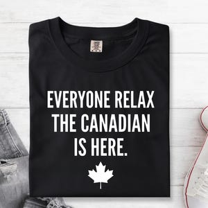 May include: A black t-shirt featuring the white text "EVERYONE RELAX THE CANADIAN IS HERE." printed in a bold, sans-serif font. Below the text is a white graphic of a maple leaf. The shirt has a crew neckline and a visible interior brand label.
