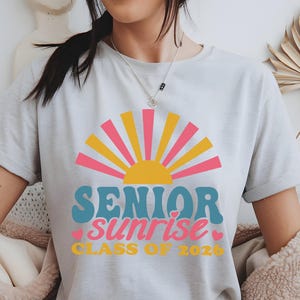 Puede incluir: Una camiseta gris claro con un gr&aacute;fico colorido. El dise&ntilde;o incluye un sol estilizado con rayos rosas, amarillos y naranjas, y las palabras "SENIOR sunrise" en azul y rosa, con "CLASS OF 2026" debajo en amarillo.