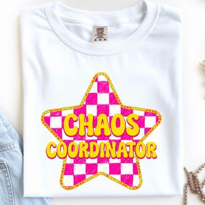 Può includere: Una t-shirt bianca con una grande grafica a forma di stella con un motivo a scacchi rosa e bianco. La stella è delineata da un design a catena dorata e contiene il testo giallo "CHAOS COORDINATOR" in un carattere grassetto e arrotondato.