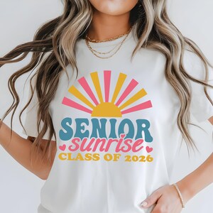 Puede incluir: Camiseta blanca con un dise&ntilde;o gr&aacute;fico colorido. El dise&ntilde;o presenta un sol de estilo retro en rosa, amarillo y naranja, con las palabras "SENIOR sunrise" en verde azulado y rosa, y "CLASS OF 2026" en amarillo.