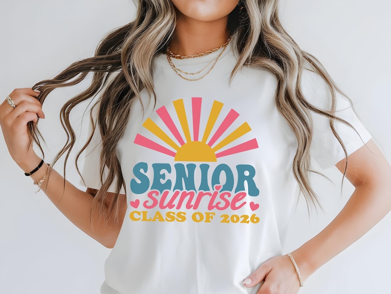 Puede incluir: Camiseta blanca con un dise&ntilde;o gr&aacute;fico colorido. El dise&ntilde;o presenta un sol de estilo retro en rosa, amarillo y naranja, con las palabras "SENIOR sunrise" en verde azulado y rosa, y "CLASS OF 2026" en amarillo.