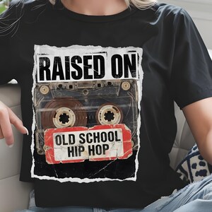 May include: Black t-shirt featuring a vintage cassette tape graphic. The design includes the text "RAISED ON" above the tape and "OLD SCHOOL HIP HOP" below, in a distressed style. The shirt is a classic crew neck.