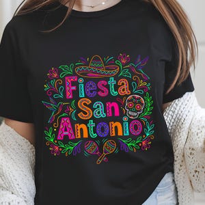 May include: Black t-shirt with a vibrant, colorful graphic that reads "Fiesta San Antonio." The design includes a sombrero, skull, maracas, hummingbirds, and floral elements in neon colors. The shirt is a crew neck.